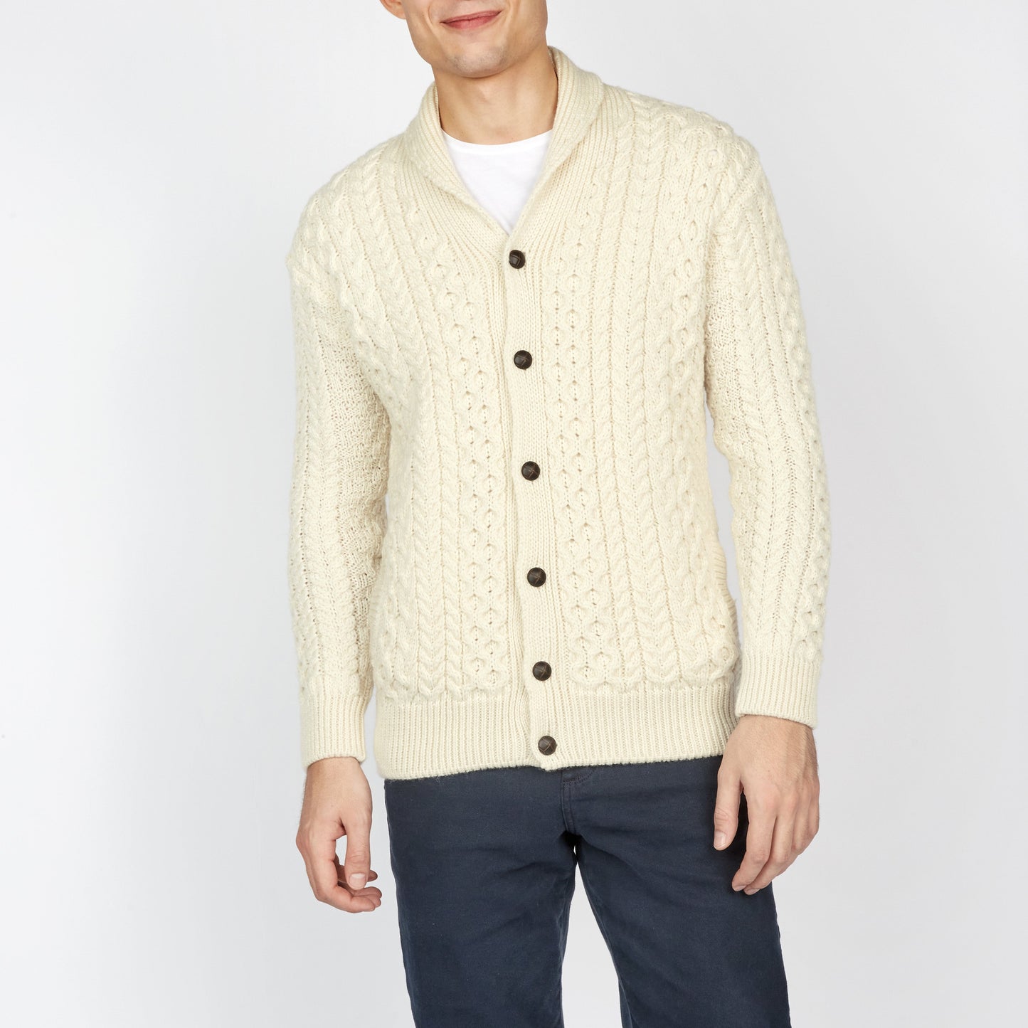 Man wearing a cream cable knit cardigan with dark pants on a white background
