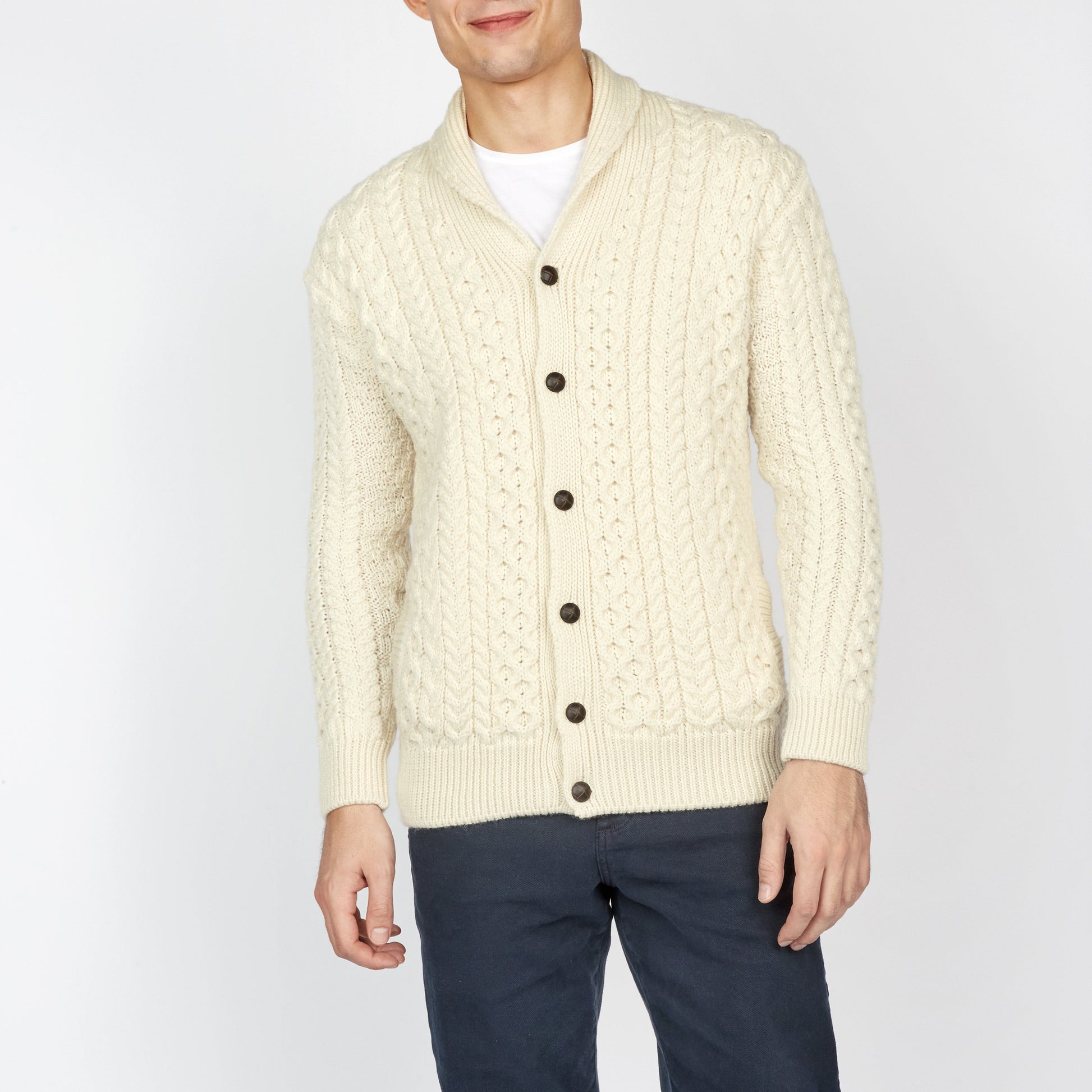Man wearing a cream cable knit cardigan with dark pants on a white background