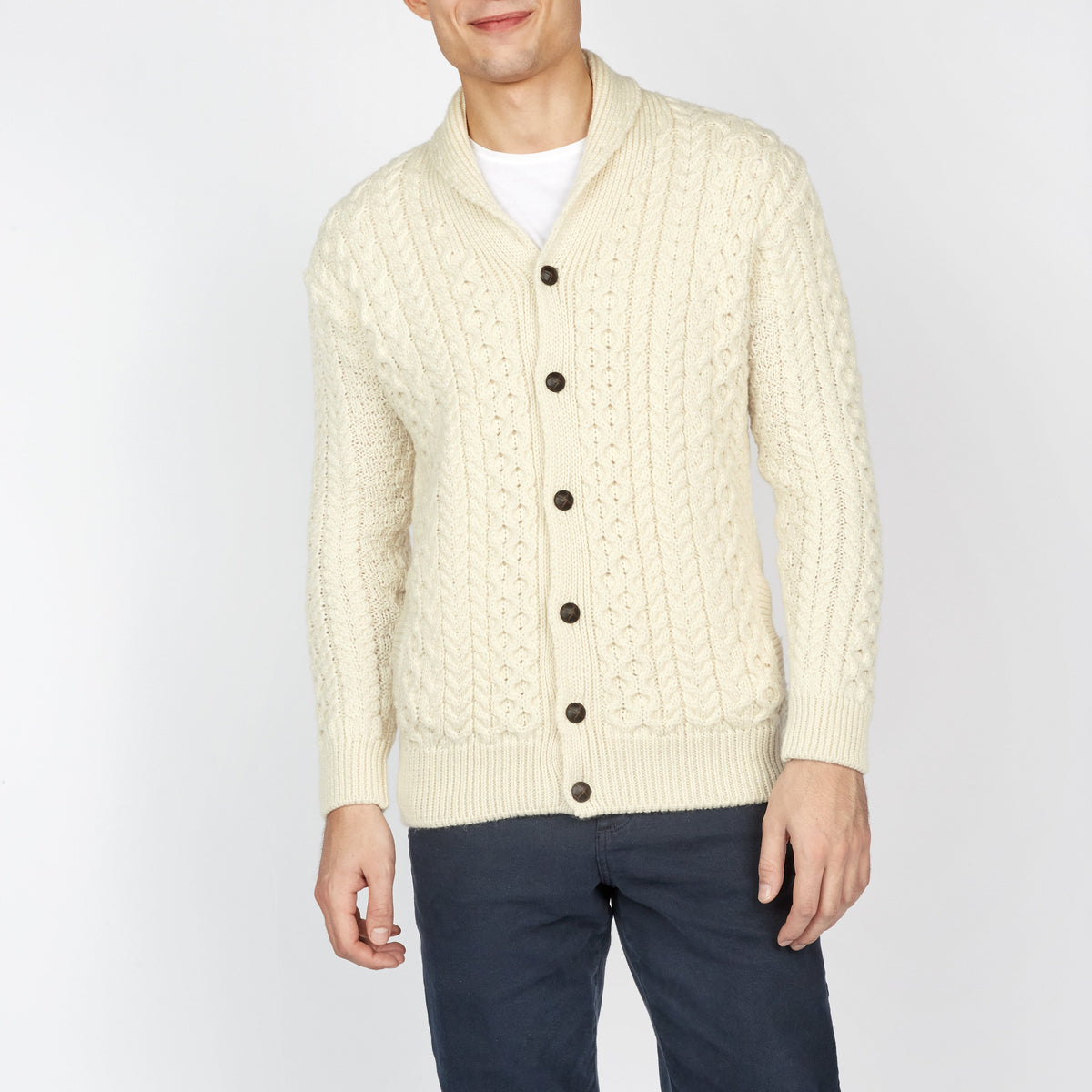 Man wearing a cream cable knit cardigan with dark pants on a white background