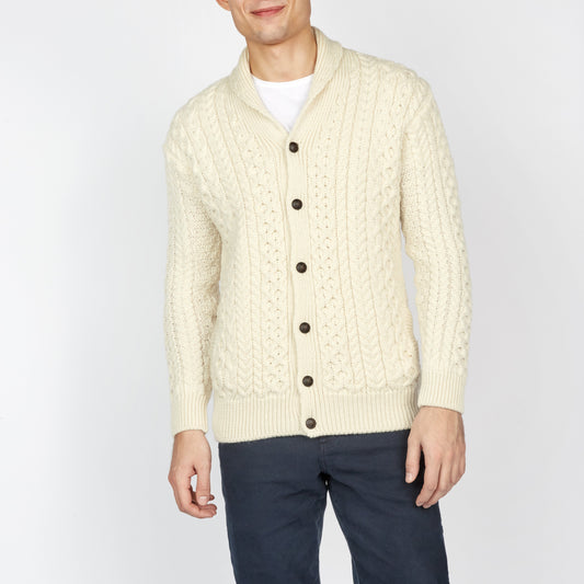 Man wearing a cream cable knit cardigan with dark pants on a white background